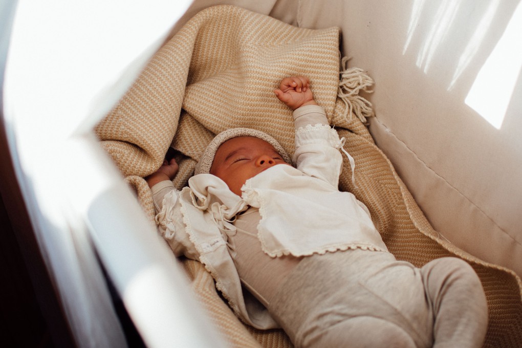 Newborn baby lying in bassinet stretching arms, clothed in white